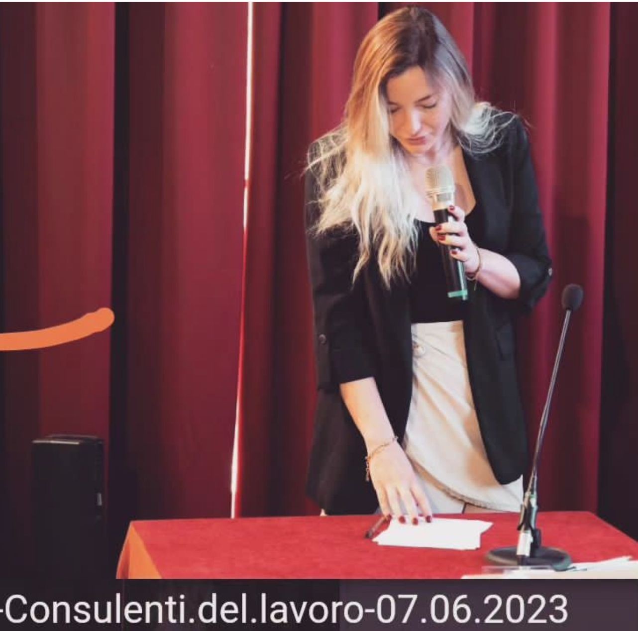 Woman with a microphone stands behind a red table and in front of red curtains.